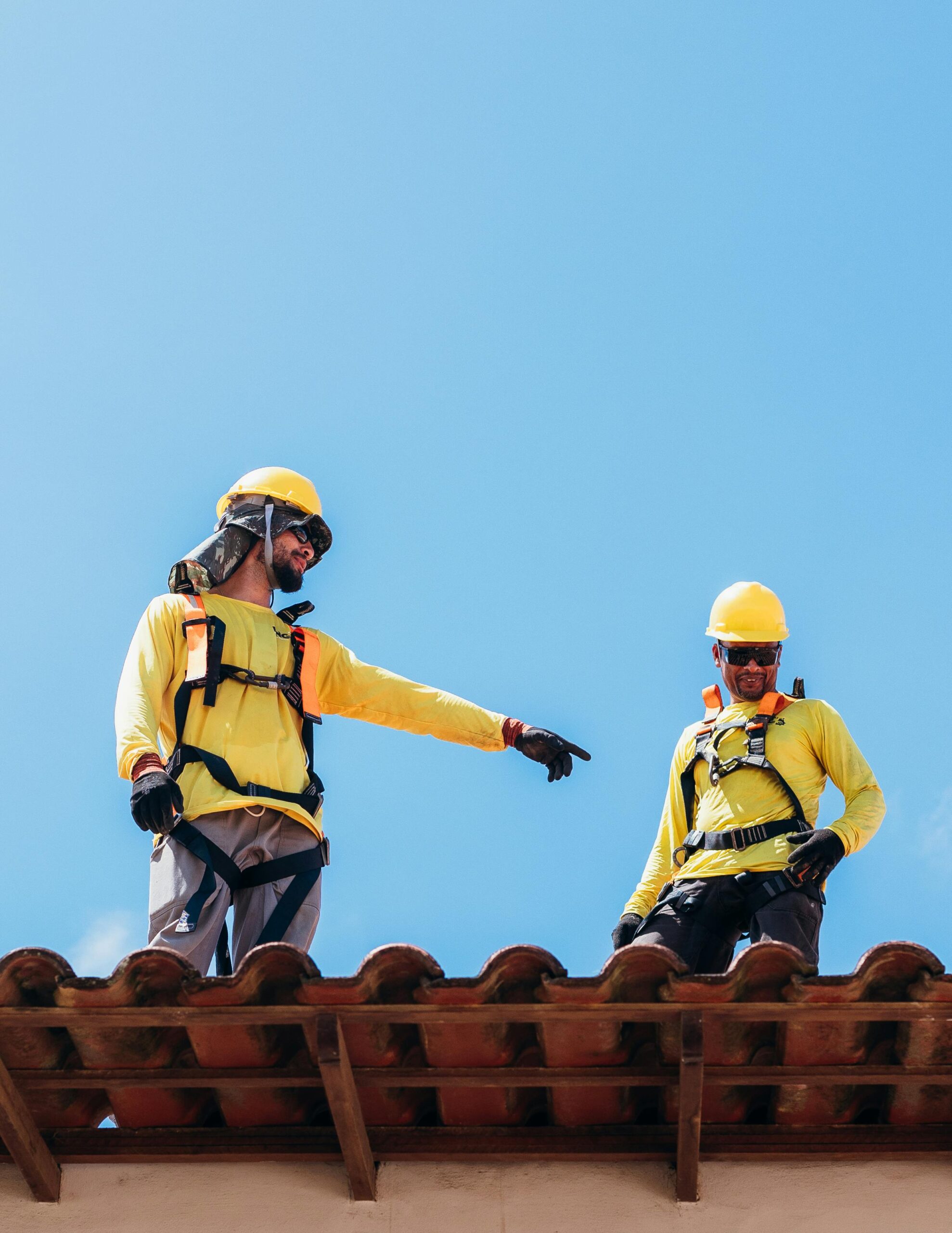 Two construction workers in safety gear discussing roofing project under clear blue sky.